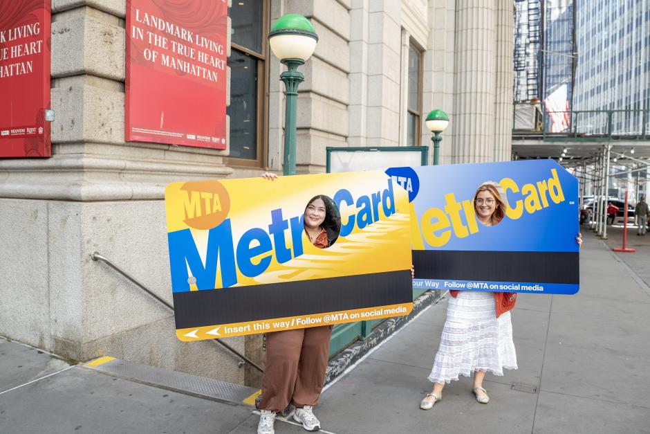 Two women smiling with oversized MetroCard posterboards outside of a subway station.