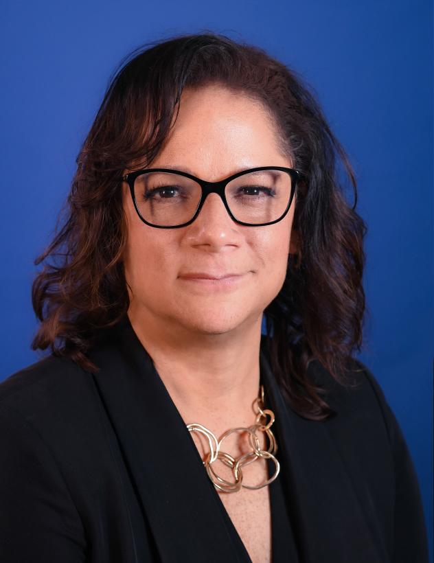 A woman with shoulder-length, curly brown hair, who is wearing a black suit jacket and a necklace. 