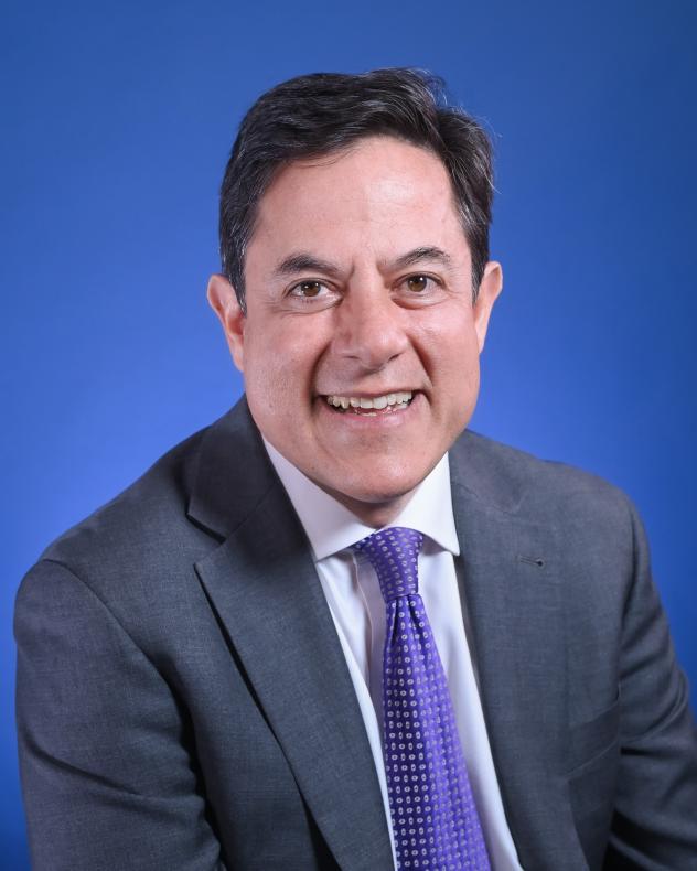 A headshot of Dan Garodnick looking at the camera wearing a dark suit, a light shirt, and a purple tie
