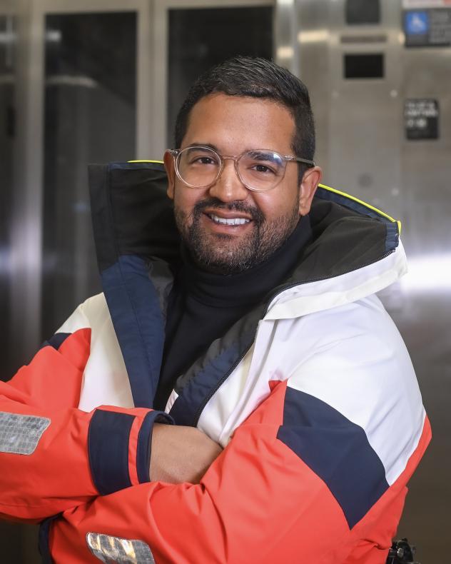 Quemuel Arroyo from the waist up wearing a brightly colored jacket with a subway elevator in the background.