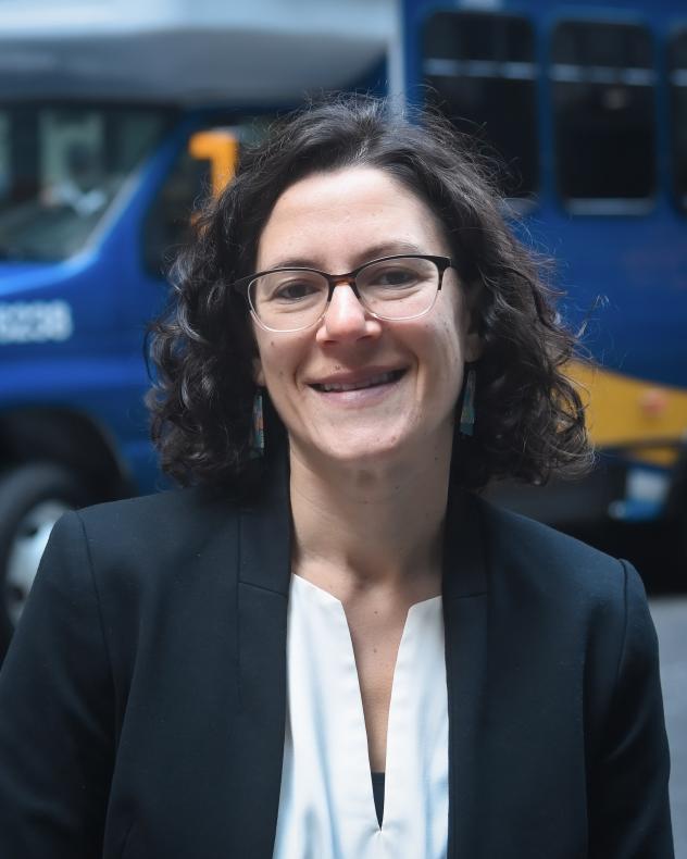 Rachel Cohen from the shoulders up wearing a dark jacket and light blouse, standing in front of an Access-a-Ride vehicle