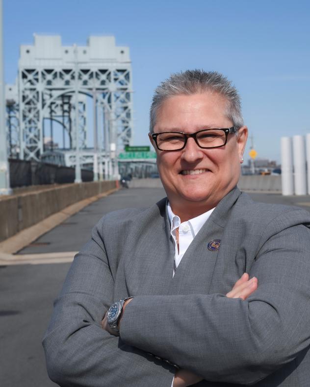 Picture of a woman with short cropped gray hair wearing a gray quarter-zip jacket in front of a bridge