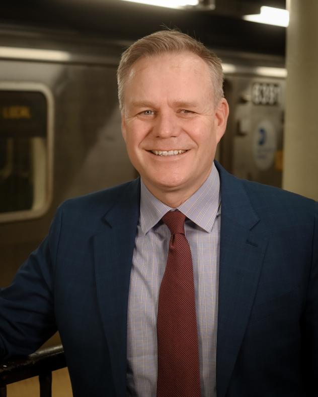 A man shot from the chest up wearing a dark suit jacket, a light shirt, and a maroon tie.