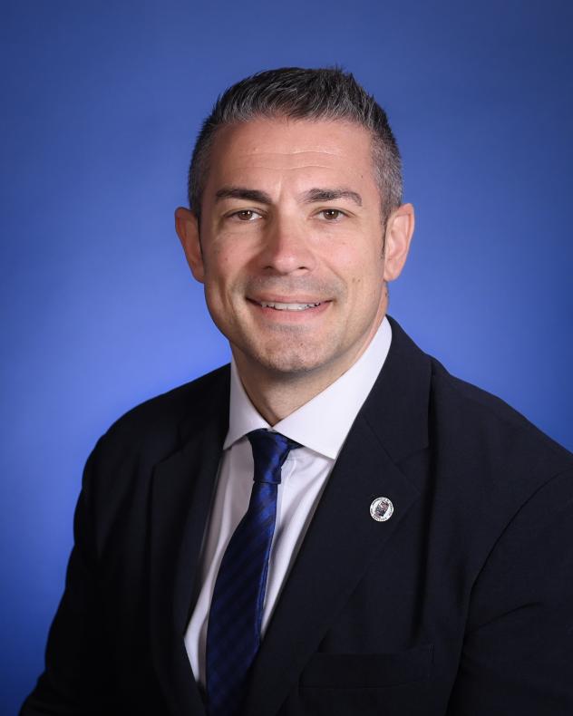 Edward Valente from the shoulders up wearing a dark blue suit, blue tie, white shirt, and ACRE lapel pin 