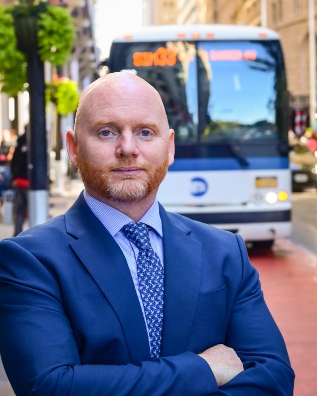 Frank Farrell in a navy blue suit jacket, blue shirt, and tie, standing in front of an express bus.
