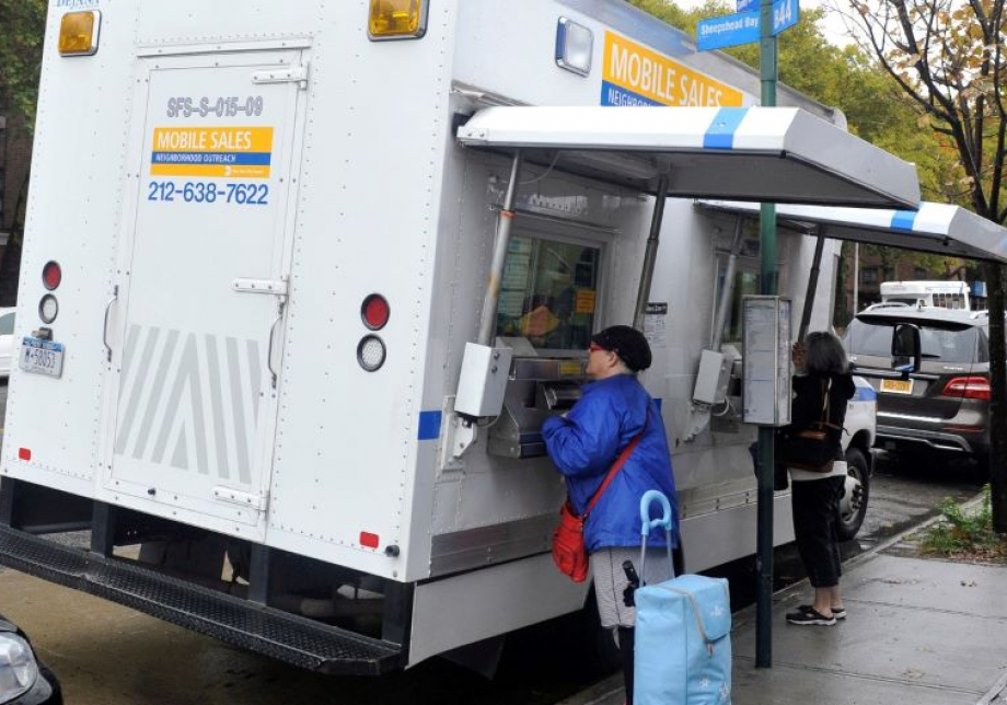 A white van with MTA branding has windows open along the side. Two people are leaning into the windows and conducting business.