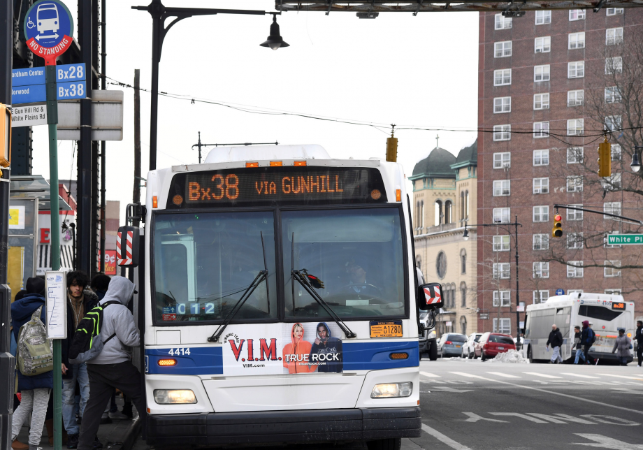 People line up to board a bus at a bus stop in the Bronx. The blue and white bus stop sign is visible in the foreground, with a bus shelter visible in the background.