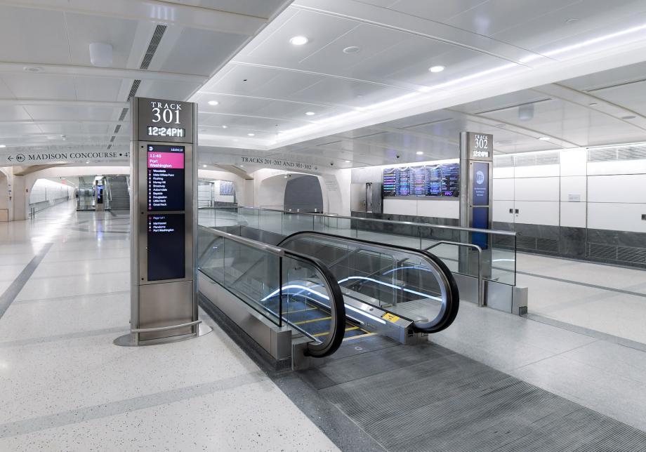 A train station with an escalator and a sign showing train times. 