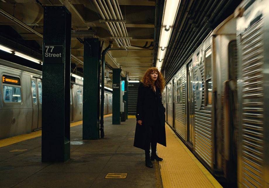 Natasha Lyonne stands on a subway platform with a train next to her 