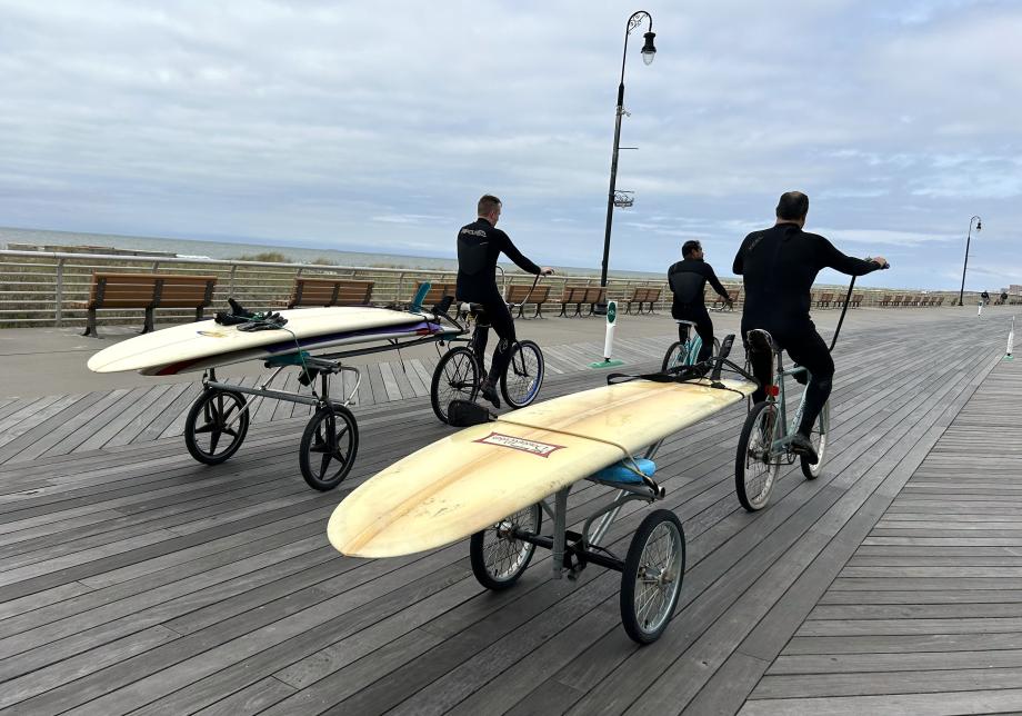Three people biking down a boardwalk with surfboards strapped to the back of their bikes. 