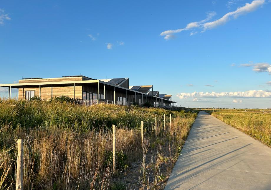 A one-story building standing next to a beach and boardwalk with grass in front of it. 