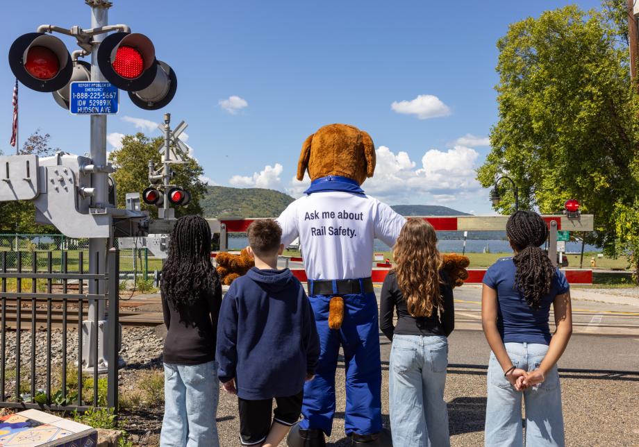 A rail-safety mascot dressed as a dog stands with four children at a railroad crossing as the gates are down and lights flash red.