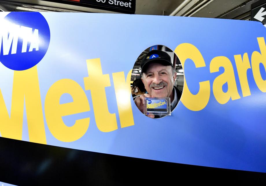 A man has his face in a hole cut out from a large blue MetroCard. He is holding a smaller blue MetroCard.