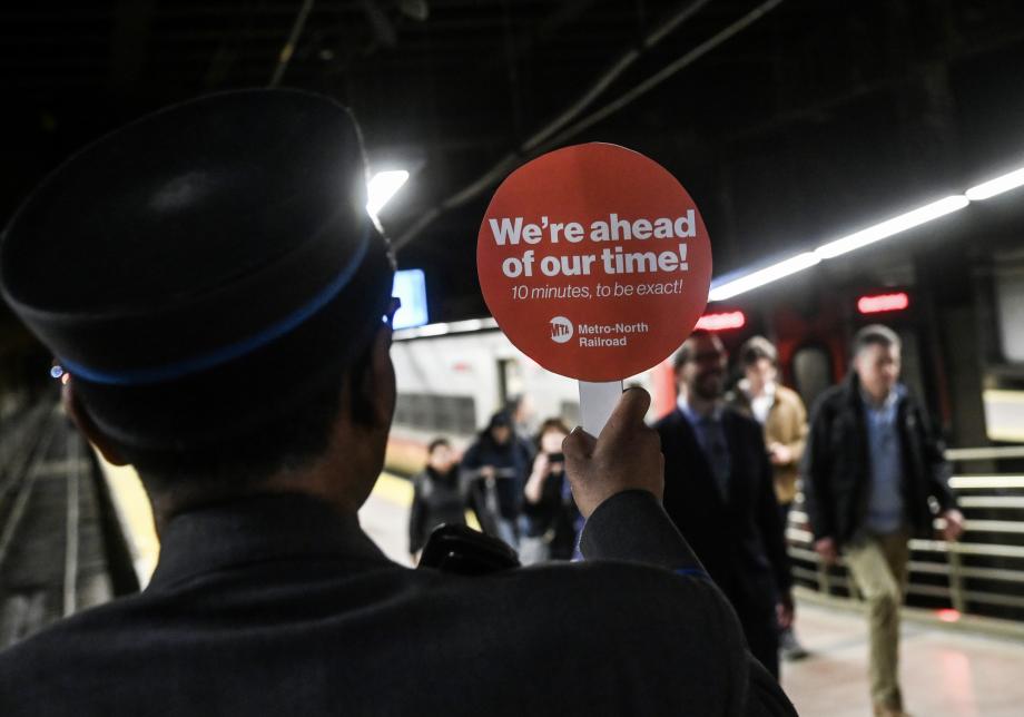 An employee on a train platform holding a lollipop sign reading "We're ahead of our time! 10 minutes, to be exact!"