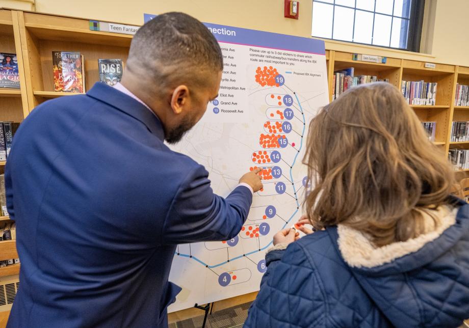 A photo showing two people looking at a large poster board showing a map of the Interborough Express route. One person is pointing at a part of the map shown on the board.