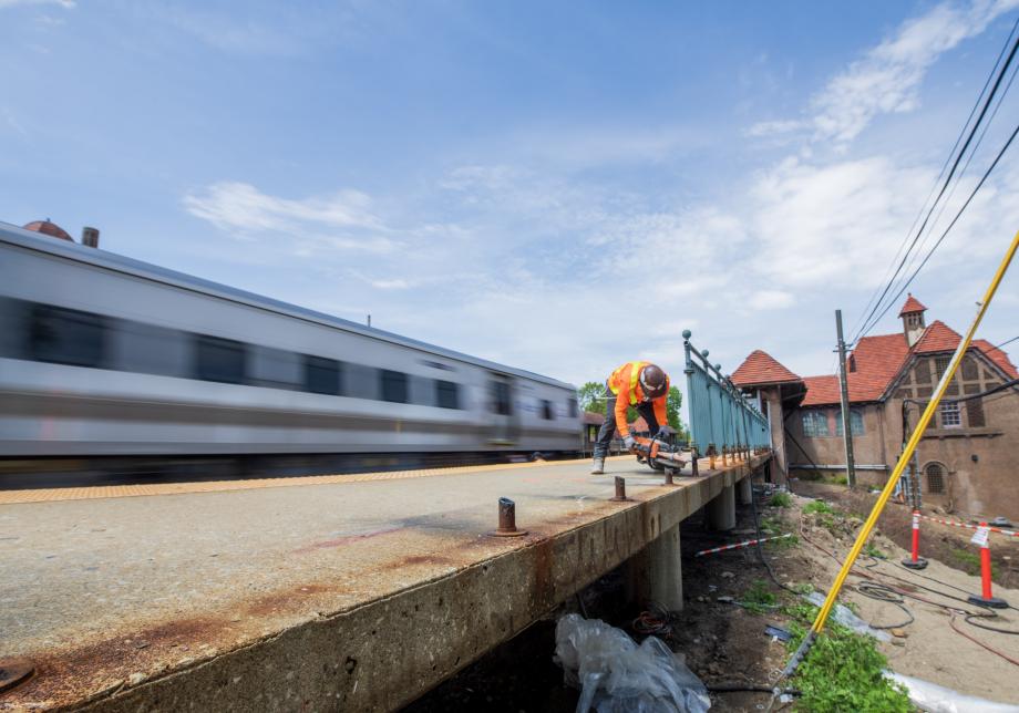 A photo showing a train rushing by the Forest Hills LIRR Station platform while a MTA worker carries out construction on the station platform. 