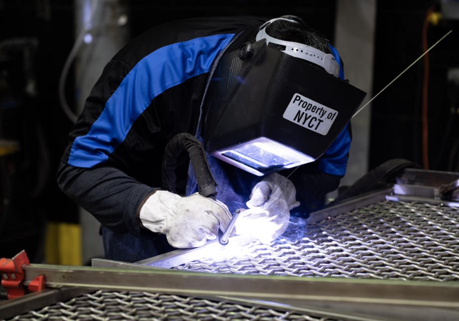 A worker in protective gear uses a welding tool to create a stainless steel platform barrier.