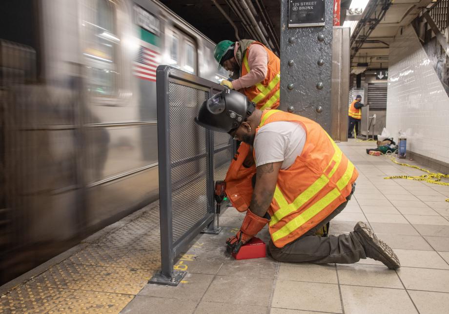 Two workers in orange vests and safety gear install a platform barrier on a subway platform.