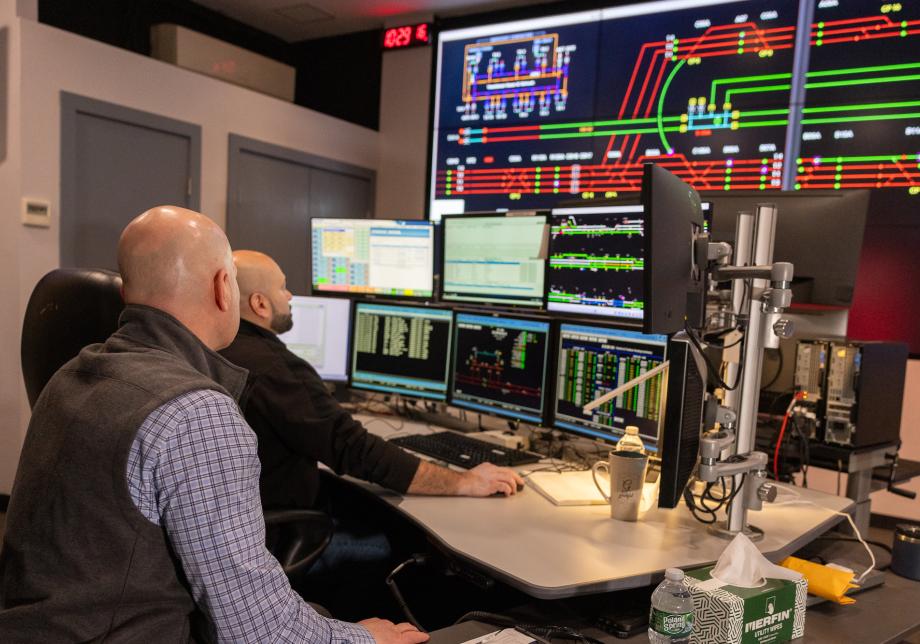 Two men sit in front of an array of screens displaying information about subway stations. 
