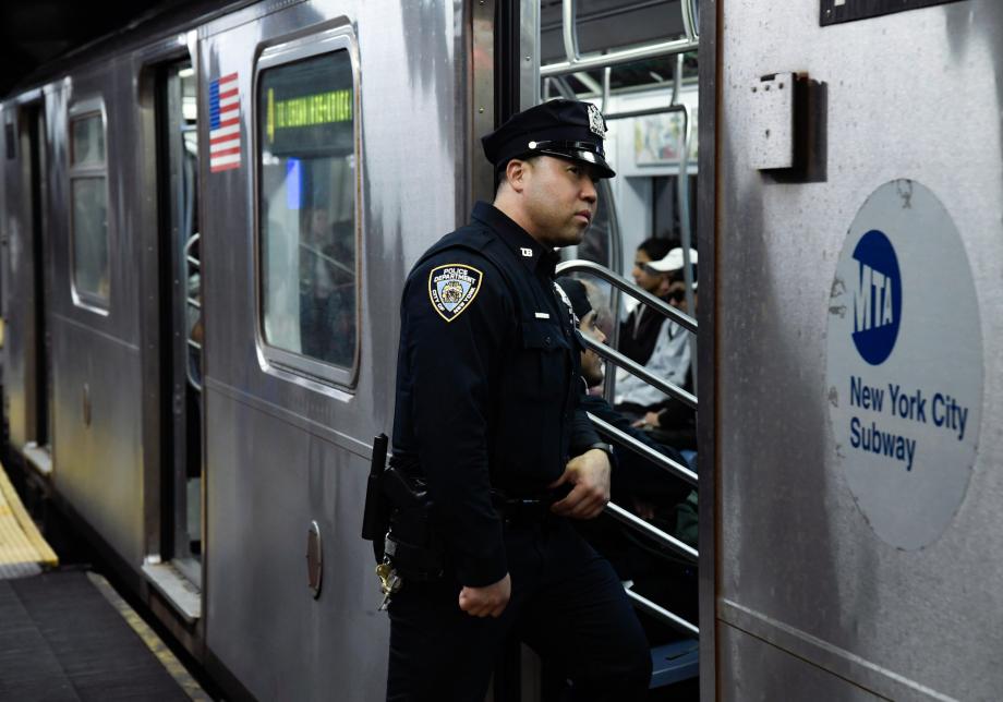 A New York City Police Department officer stands in the doorway of a subway train. He is looking inside the subway car. 