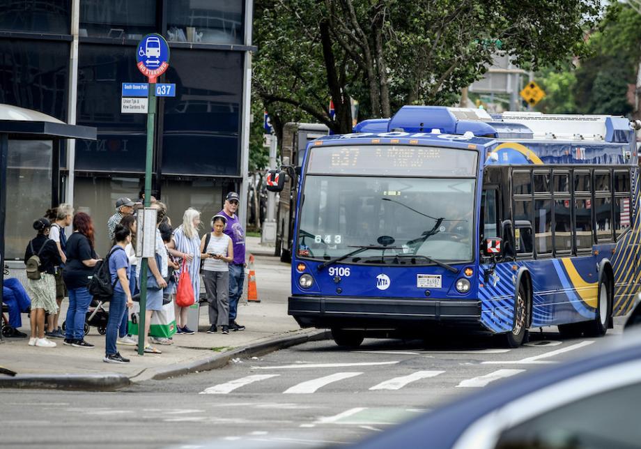 A group of people waits at a bus stop, as a bus pulls up to the curb in front of them.