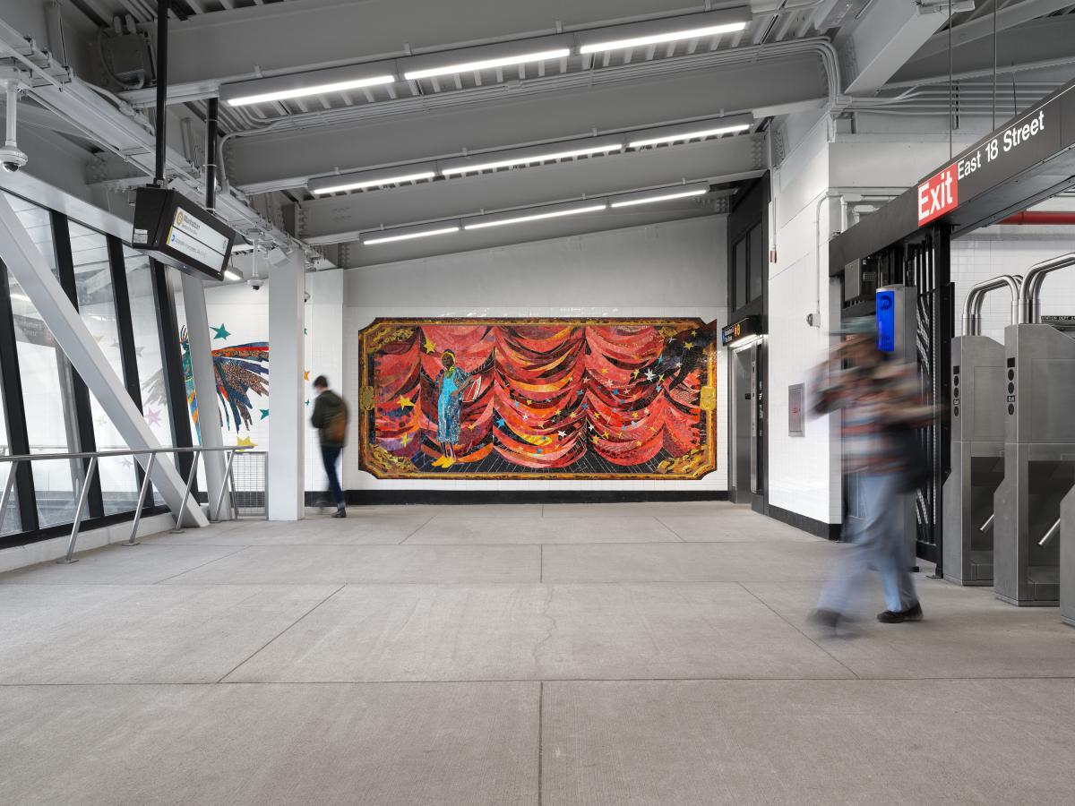 Subway station with a colorful mosaic mural depicting a red curtain and figures. Two blurred pedestrians walk past.