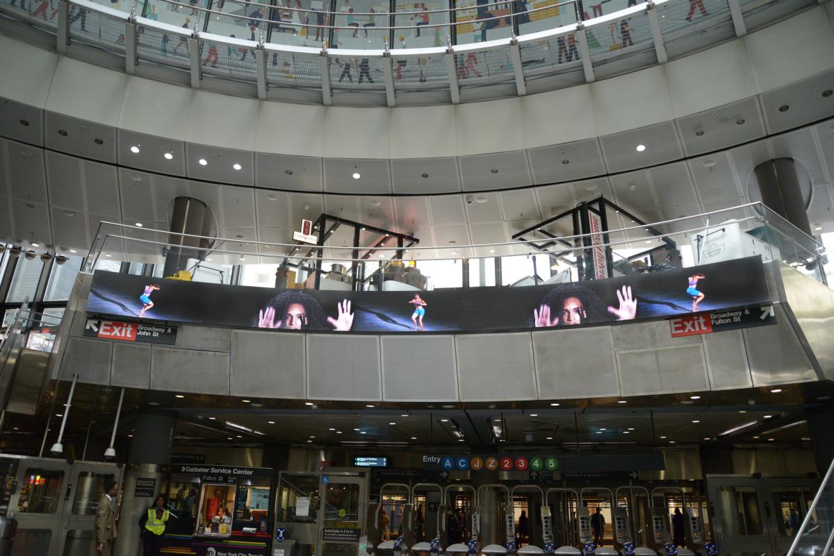 Interior of Fulton St Station with a curved LED screen displaying a woman's face and dancers. Below are turnstiles and signage for the A, C, 2, 3, 4, and 5 trains.