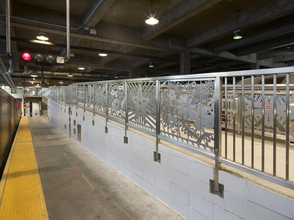 Train platform with concrete flooring, yellow safety strip, and decorative metal railings featuring floral patterns, swimming fish, and stars. Train signals can be seen overhead.