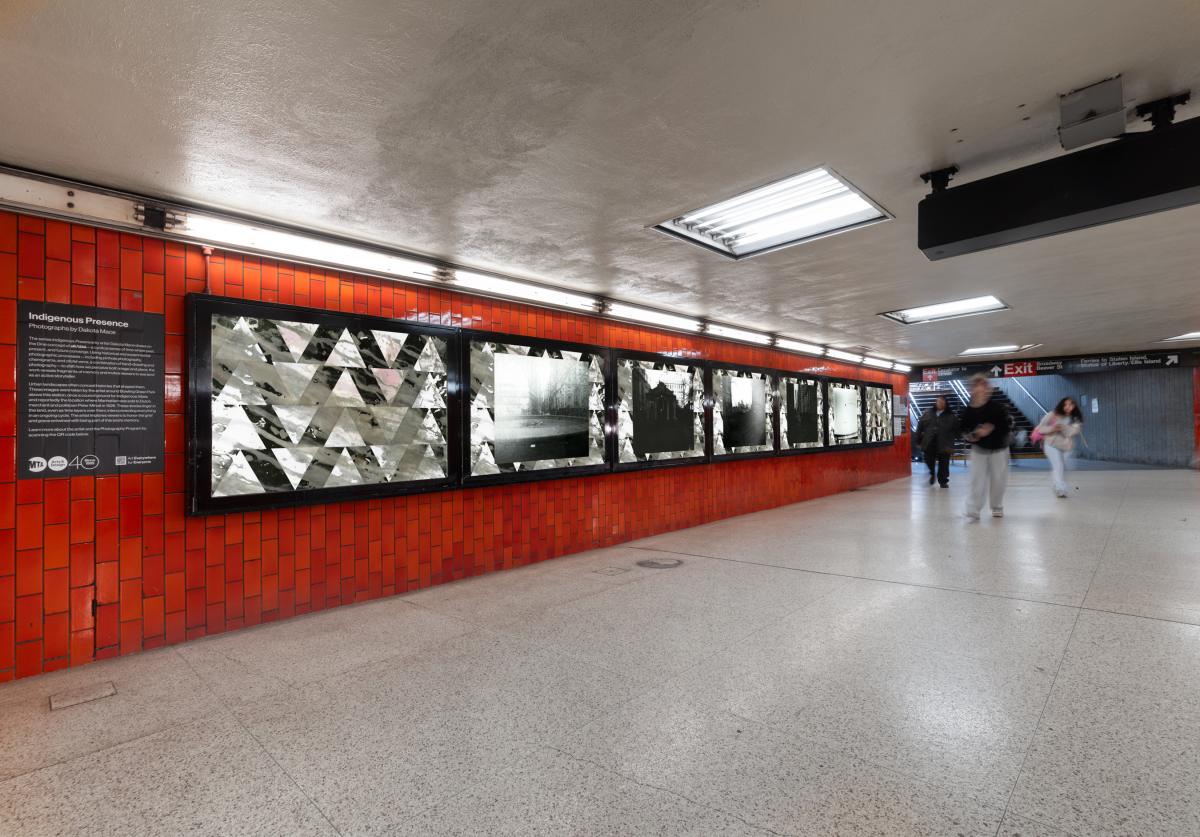 Subway station with red tile walls displaying a series of black and white photos. People walk past in the foreground.