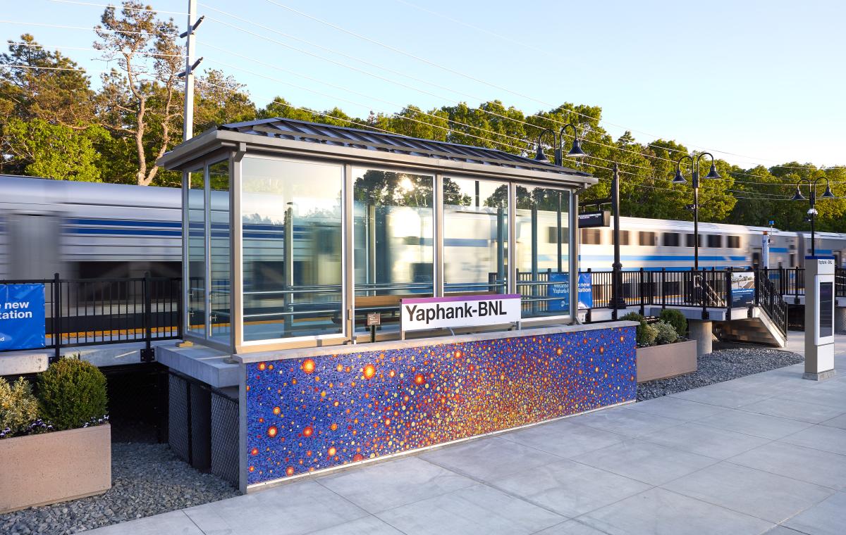 Yaphank-BNL train station with a glass shelter decorated in colorful mosaic. There is a train passing in the background.