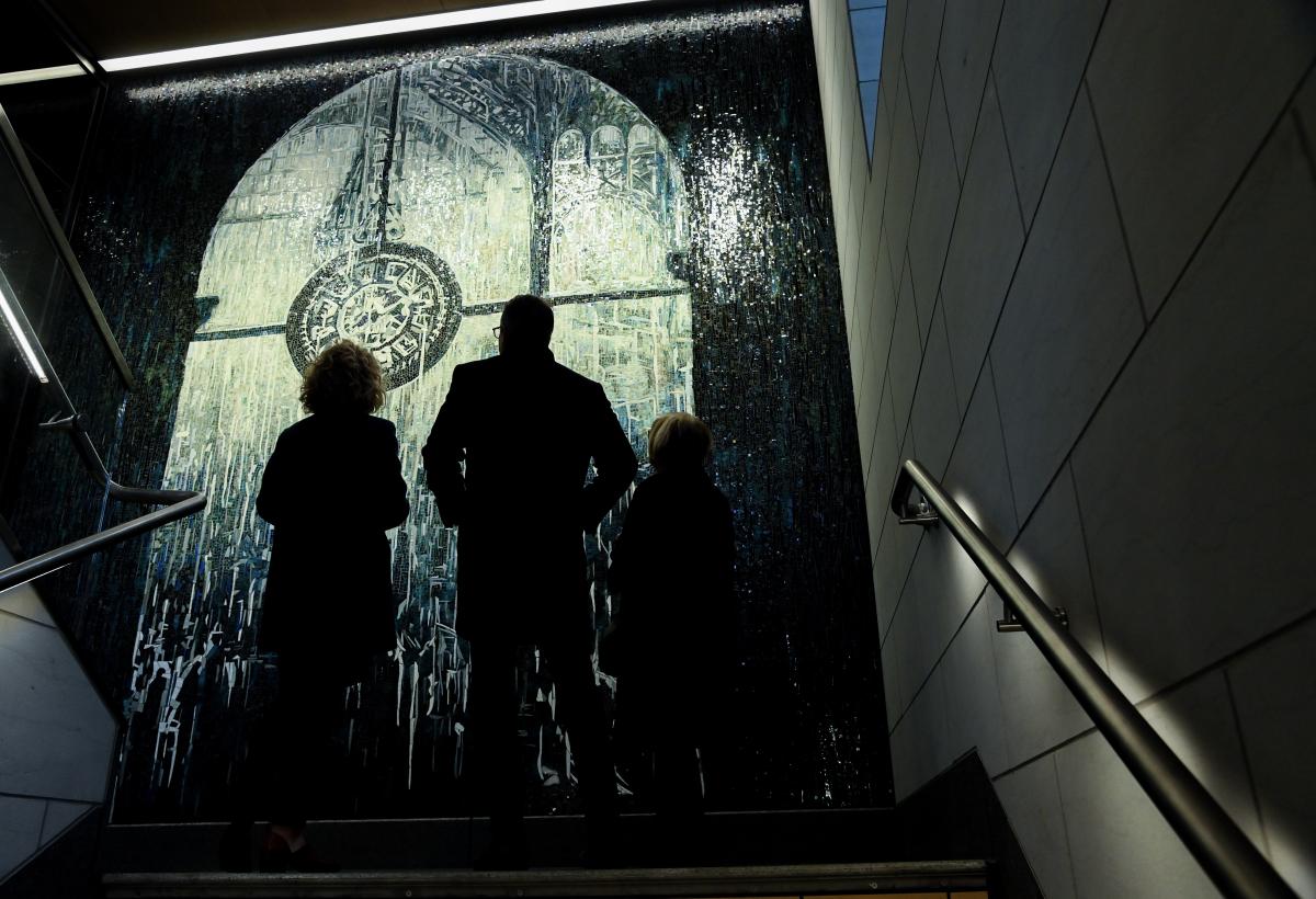 A group of three people stands in front of a large mosaic artwork that depicts a clock, which is illuminated by overhead light. They are silhouetted against the mosaic.
