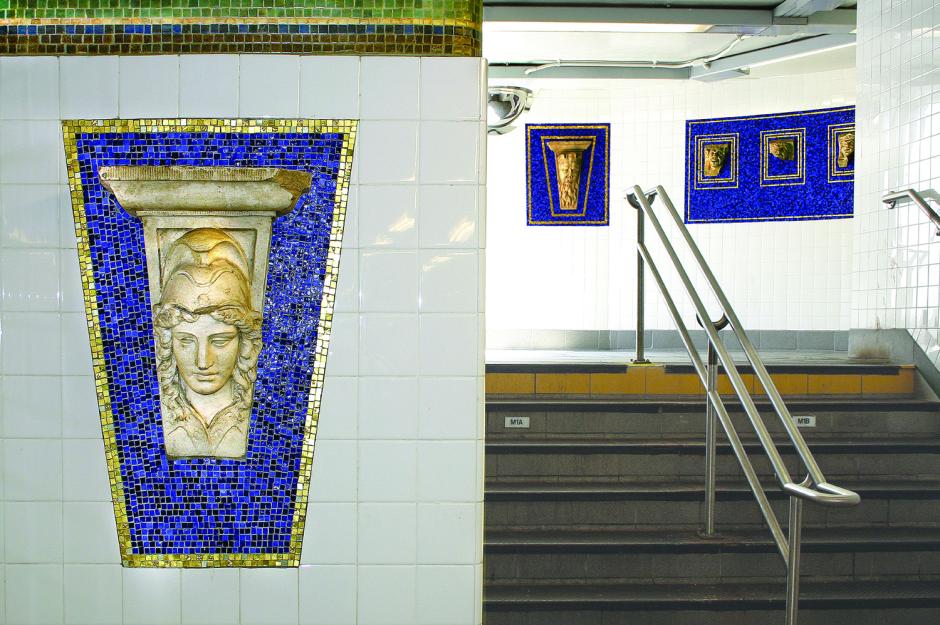 Artwork in terra cotta artifacts by the Arts for Transit Collaborative showing ornamental architectural forms salvaged from buildings and glass mosaic designs surrounding them on mezzanine and stairway walls. 