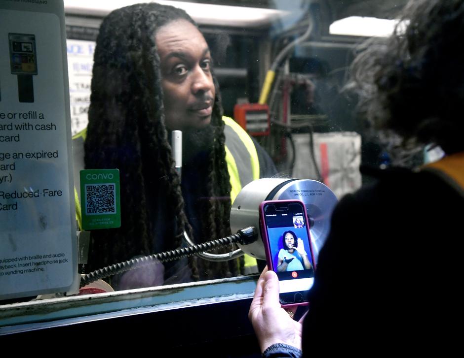 A customer standing at a ticket booth speaking to an agent, holding a smartphone with a sign language interpreter on the screen