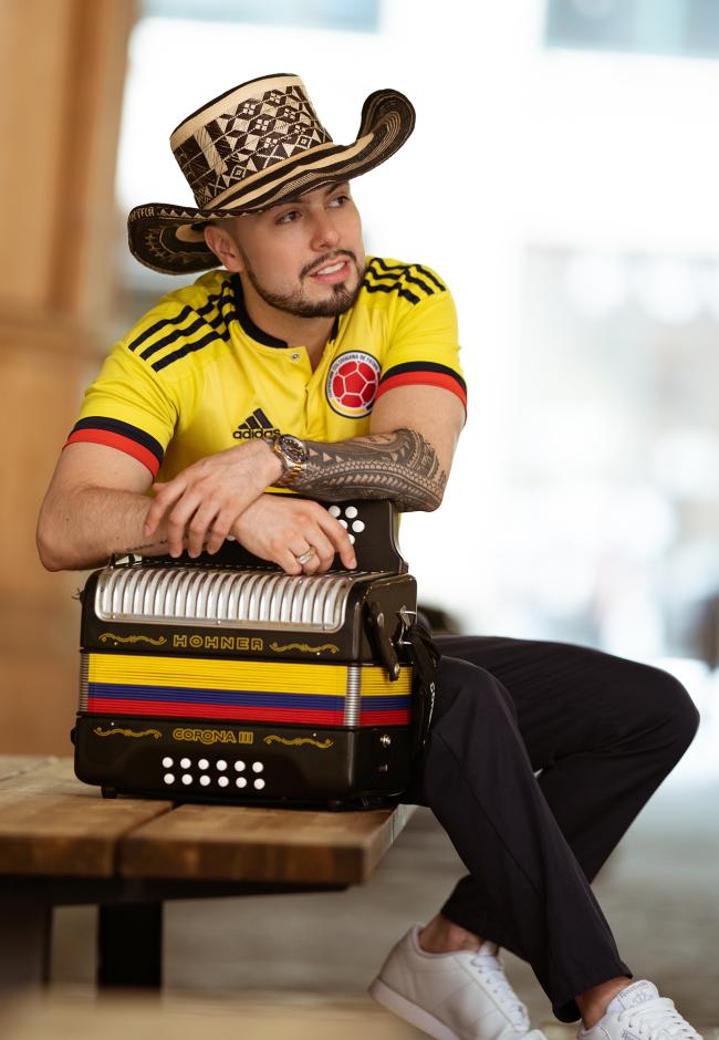 Man sitting on a bench wearing a Colombian hat and jersey holding an Accordion.