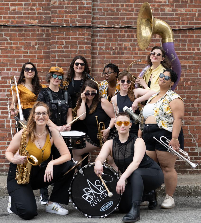 Ten women dressed in black and gold wearing sun glasses posing with various brass and percussion instruments in front of a red brick wall.