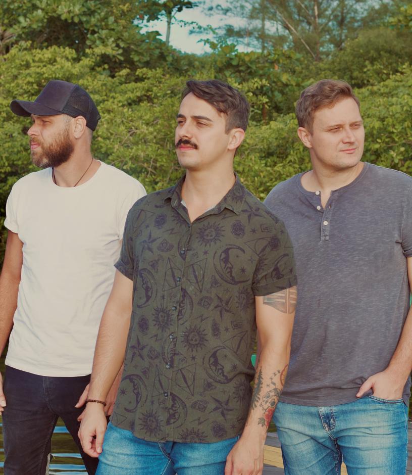 Three men standing in front of trees. One in a hat, two with facial hair. One wearing white shirt, one in grey shirt, and one in moon pattern shirt.