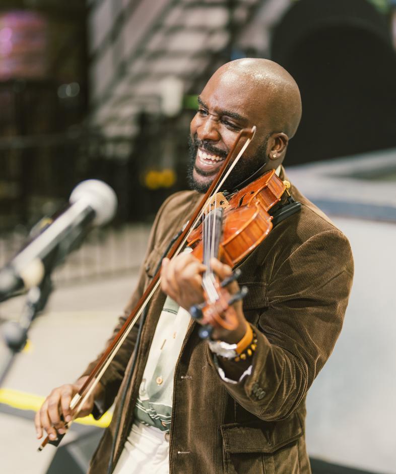 Man in brown jacket playing violin in front of a blurred background.