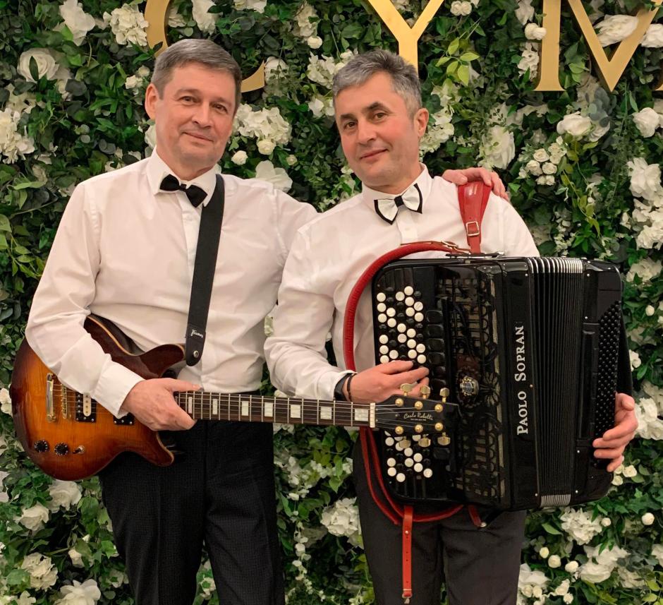 Two men in front of flower and vine wall wearing bow ties and holding a Guitar and an Accordion.