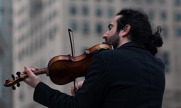 Man with pony tail and facial hair playing Violin in front of tall building with his back to the camera.