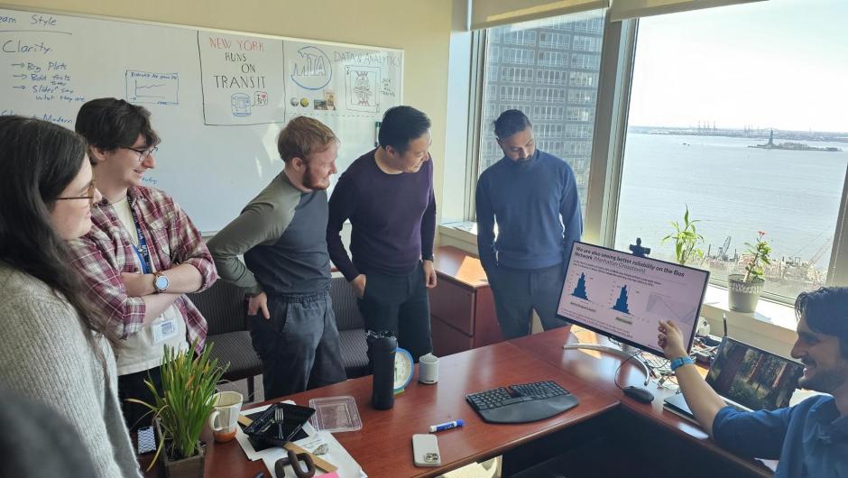 Five people gathered around a monitor. A sixth person is sitting behind a desk pointing at the monitor. A large window looking out on the Statue of Liberty is in the background.