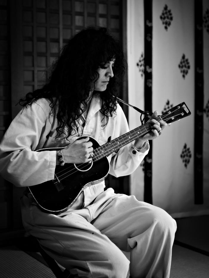 Black and White photo of woman in front of white cloth with flower pattern and lattice wood wall holding a Ukulele.