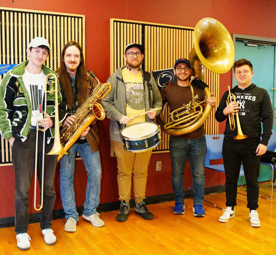 Five men dressed in different jeans and jackets holding brass and percussion in a room with wood floors and paneled walls.