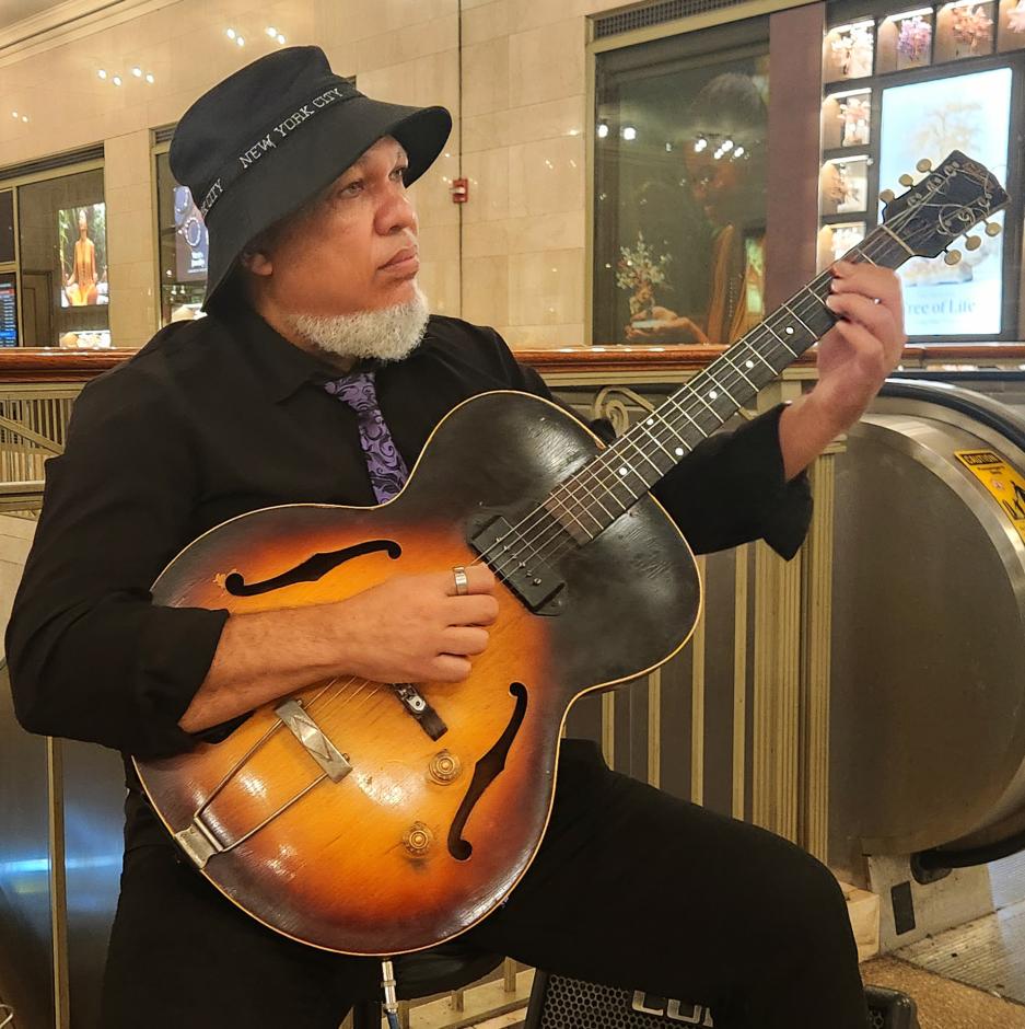 Man dressed in all black with hat and purple tie playing guitar in front of escalator.