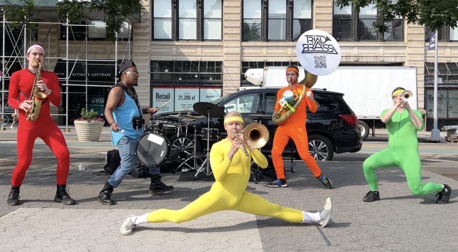 Five men dressed in different color leotards playing brass and percussion on the sidewalk next to a tree and black truck.