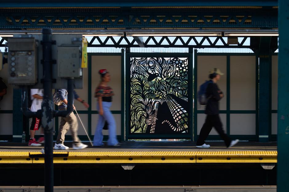 People walking by a metal artwork in a station platform windscreen.