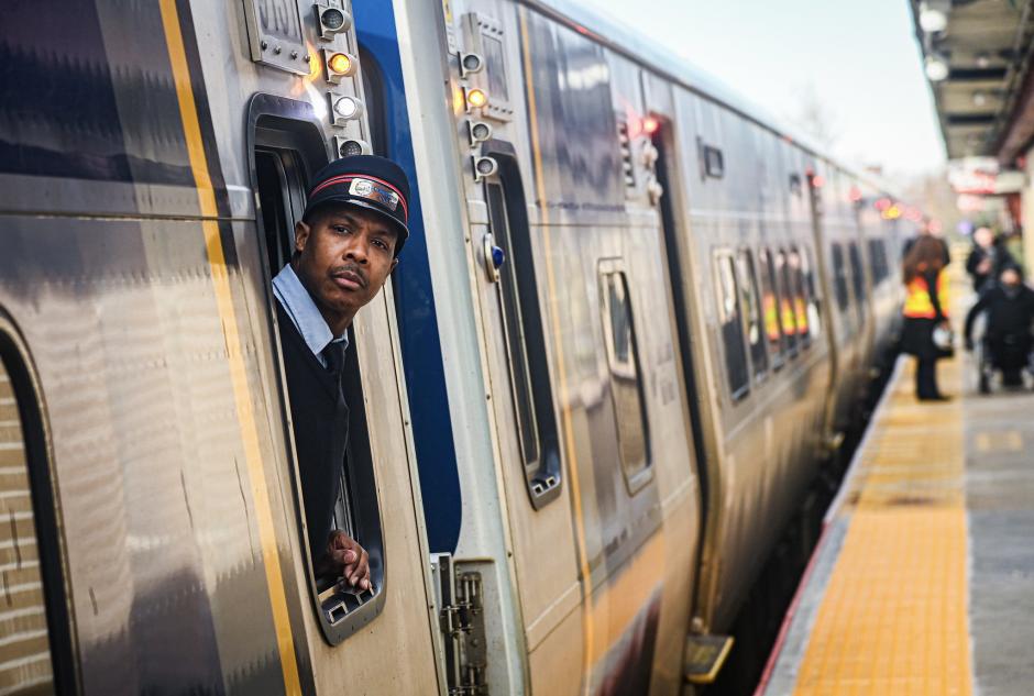 An LIRR M9 train at Laurelton Station, with the conductor looking at the camera out the window