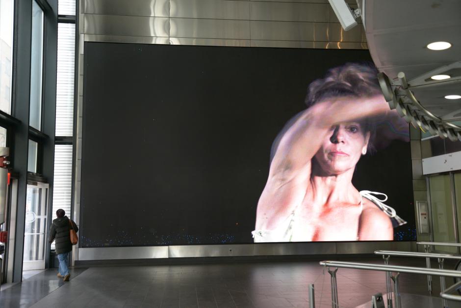 A large indoor video screen shows a person with an intense expression, arm raised.