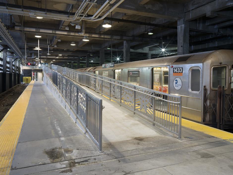 Inside asubway station, an empty trainis parked beside a intricately detailed metal railing on a platform ramp. Yellow tactile paving lines the platform edge.