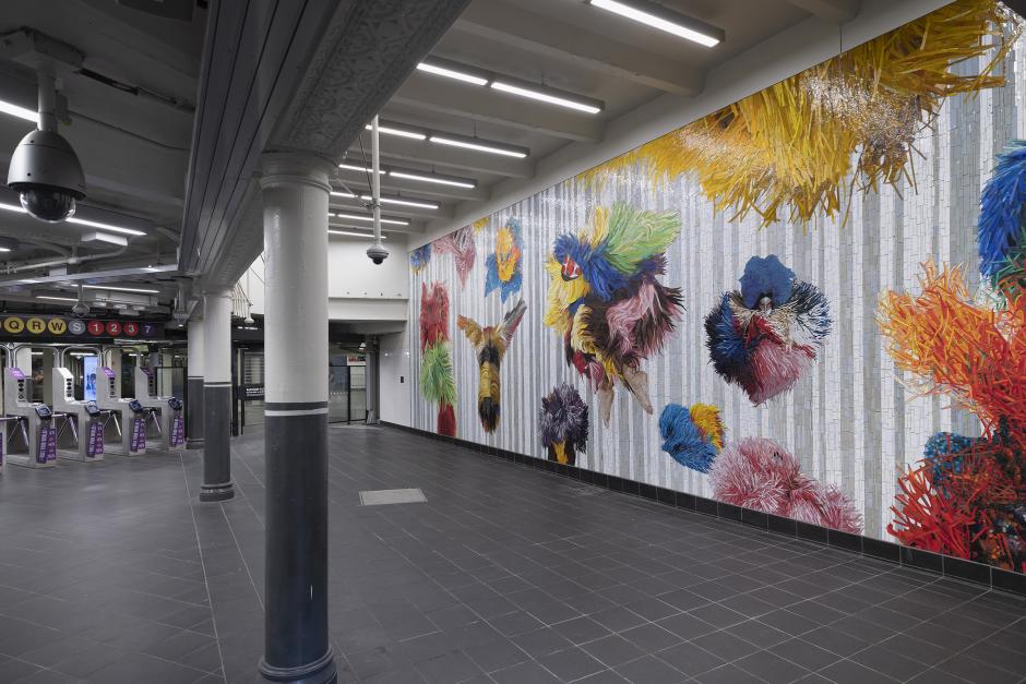 A vibrant mosaic artwork located within a subway station, extending across a large wall, featuring an array of abstract, fluffy, and textured shapes, primarily in bright colors such as yellow, red, blue, and green. The mosaic elements are arranged against a backdrop of vertical silver and white stripes.