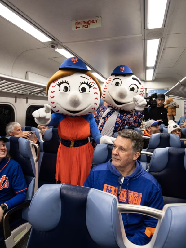 Mr. and Mrs. Met stand in the middle of a Long Island Rail Road train car with Mets fans wearing jerseys sitting in the seats